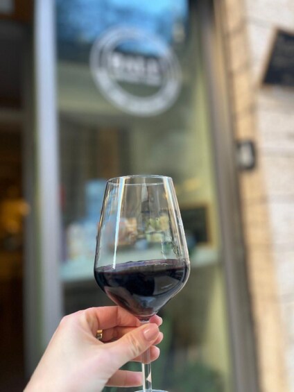 A hand holding a wine glass with red wine, in front of a building with a sign. 3