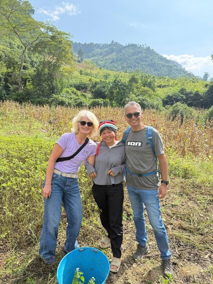 Three people standing in a field with a blue bucket and plants in the foreground, surrounded by greenery and hills in the background. 4