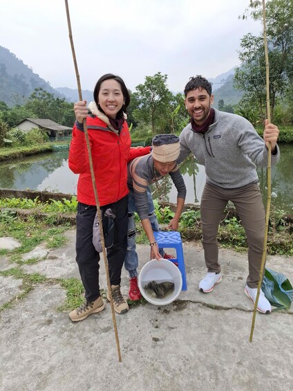 Three people near a body of water, one holding a stick, another bending over a bucket, and a third standing with a stick. 2