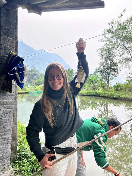 A person holding a fish, another person fishing, a river, and greenery in the background. 5