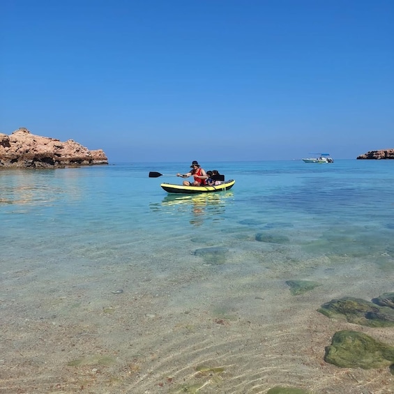 A clear blue sea with a kayak carrying two people and a child, a rocky shore, and a distant boat. 2