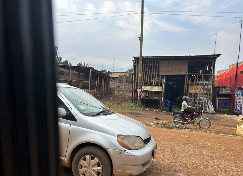 A silver car parked on a dirt road, a motorcycle with a rider, and a small shop with a sign reading 'Charcoal from Congo'. 3
