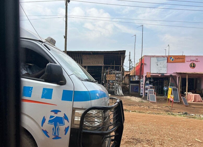 A white van with blue stripes and recycling symbols parked on a dirt road. In the background, there are buildings with signs, including one for a shop named 'Starlines' and another with a sign that reads 'CHARGES FROM CONGO'. 5