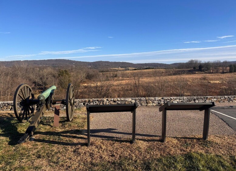 A cannon, a stone wall, and a bench in a rural landscape with hills in the background. 1