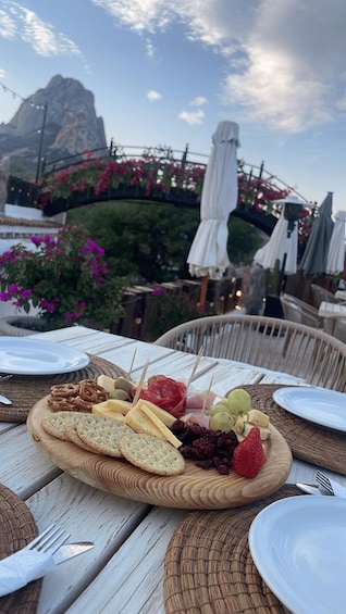 A table with a charcuterie board, wicker chairs, and umbrellas in a garden with a mountain backdrop. 2