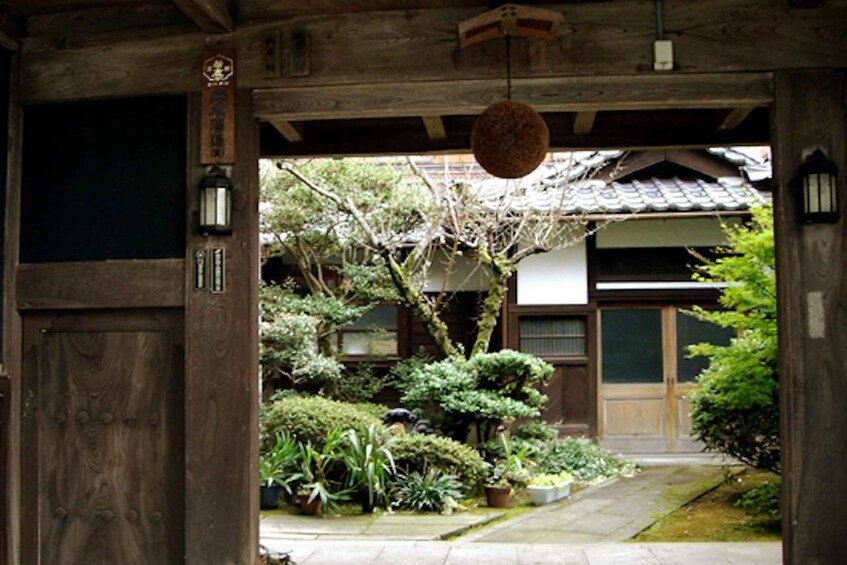 A traditional Japanese wooden gate with a hanging lantern, a well-maintained garden with various plants, and a building with a tiled roof in the background. 1