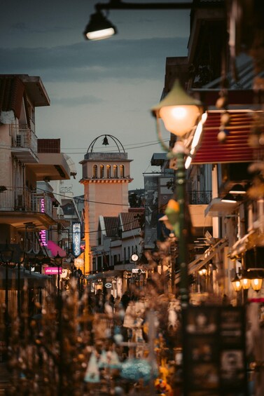 A bustling street at dusk with traditional architecture, including a tower with a dome and arches, and various shops with illuminated signs. 5