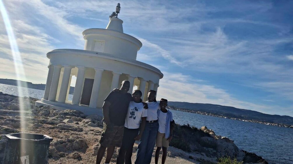 A group of people standing in front of a white lighthouse with a statue on top, overlooking a body of water and a distant coastline. 3