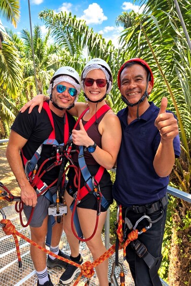 Three people wearing harnesses and helmets, standing on a metal platform with ropes, surrounded by palm trees. 3