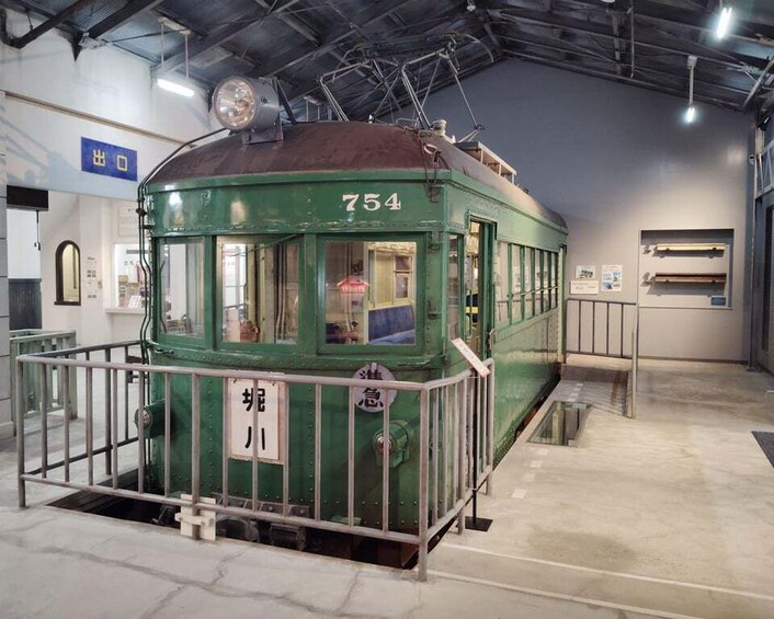 A green tram with the number 754, enclosed by a metal fence, in an indoor setting with a high ceiling and industrial lighting. 4