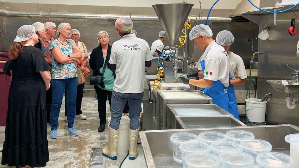 A group of people in a kitchen, some wearing hairnets and aprons, with a large industrial mixer and containers in the background. 1