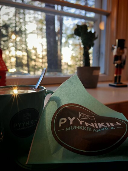 A cup with a spoon, a green napkin, and a window with a view of trees and a plant. 1