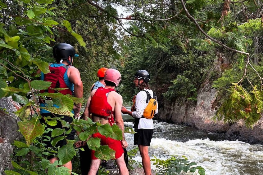 Three people wearing helmets and life jackets standing by a flowing river surrounded by dense greenery. 2