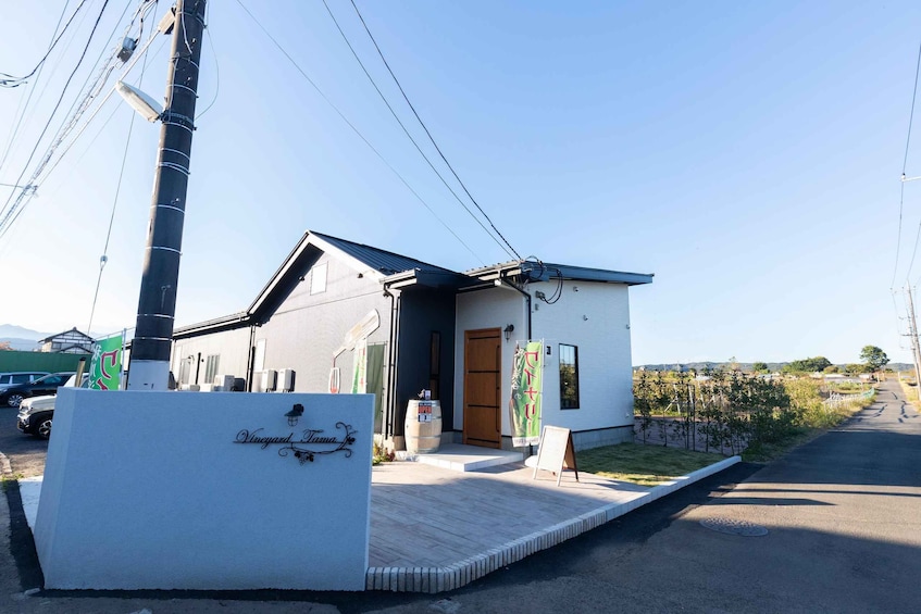 A small white building with a dark roof, a wooden door, and a sign that reads 'Vineyard Tama.' There are power lines above and a paved area in front. 1