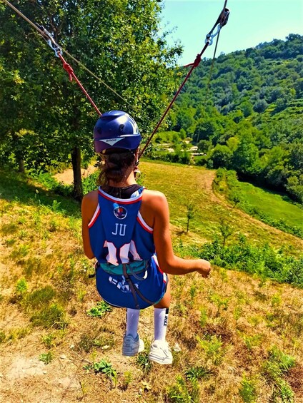 A person on a zip line with a blue helmet and a numbered jersey, overlooking a green landscape. 1