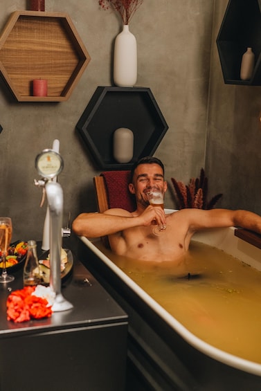 A person relaxing in a bathtub with a towel wrapped around their neck, surrounded by various toiletries and a glass of beer. 2