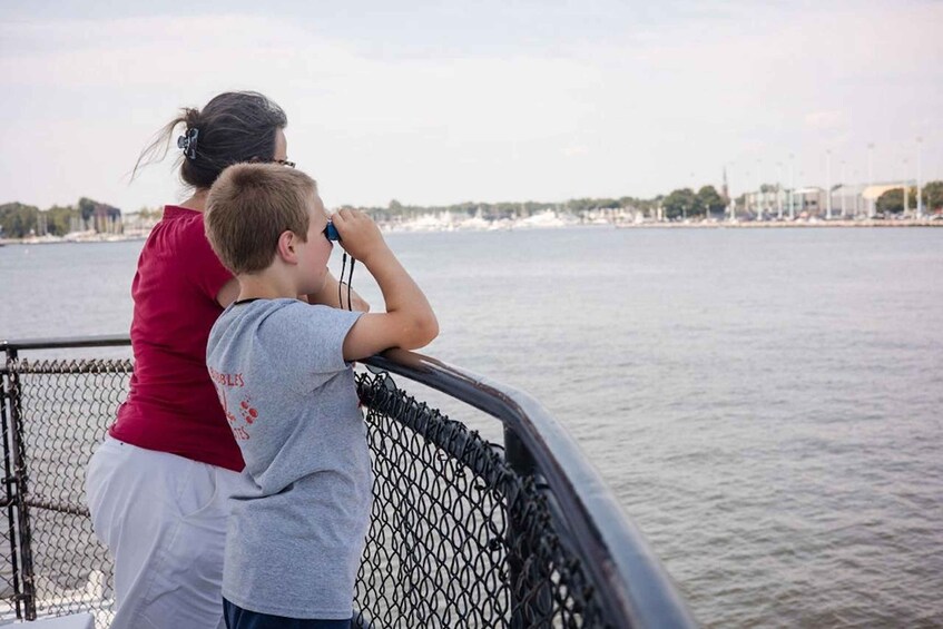 A person in a red shirt and white pants standing next to a child on a dock, looking out over a body of water with a cityscape in the background. 1