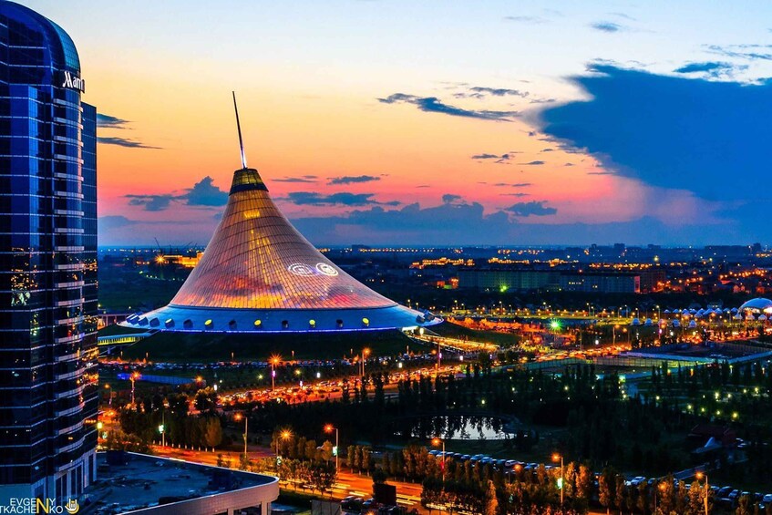 A cityscape at dusk with a prominent conical building with illuminated patterns, surrounded by modern high-rises and a busy street with cars. 1