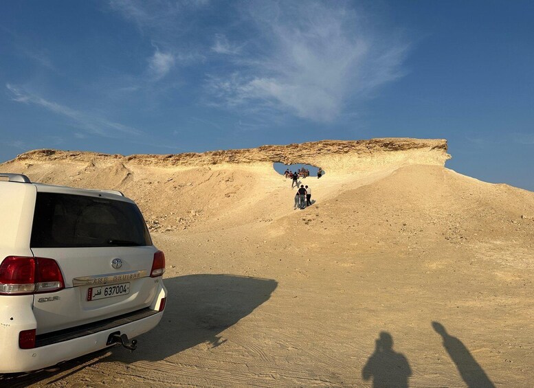 A white SUV parked on a sandy terrain with a group of people ascending a rocky hill. 2