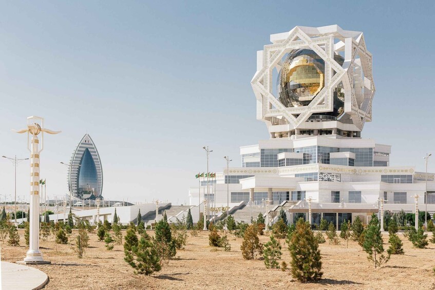 A modern architectural complex with a distinctive tower featuring a spherical element, surrounded by smaller buildings and a landscaped area with young trees. 1