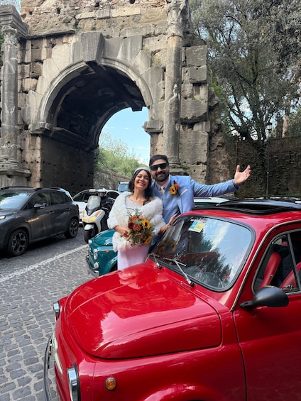 A couple posing in front of a red vintage car with a stone archway in the background. 2