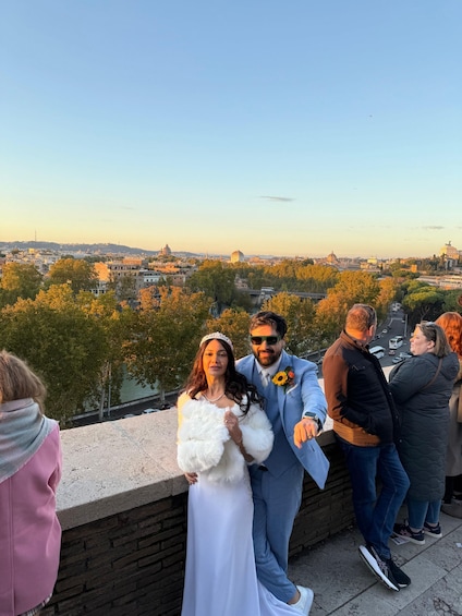 A couple in wedding attire on a balcony overlooking a cityscape with historic buildings and trees. 3