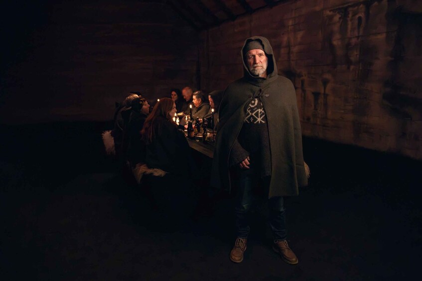 A person standing in a dimly lit room with wooden walls, a table with bottles, and a group of people in the background. 2