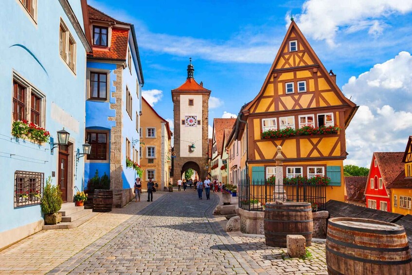 A cobblestone street lined with colorful half-timbered houses, a central tower, and pedestrians. 1