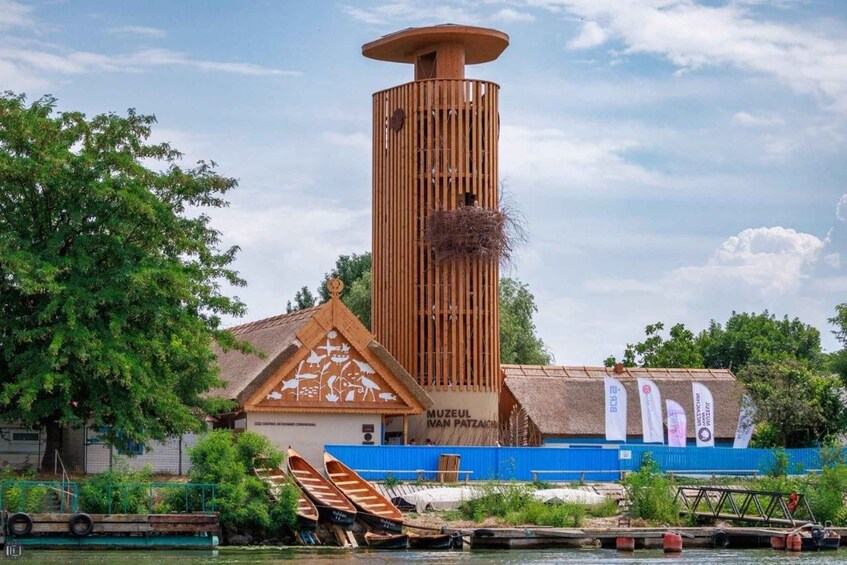 A museum with a tall wooden tower, a traditional building with a thatched roof, and several boats docked by the water. 1