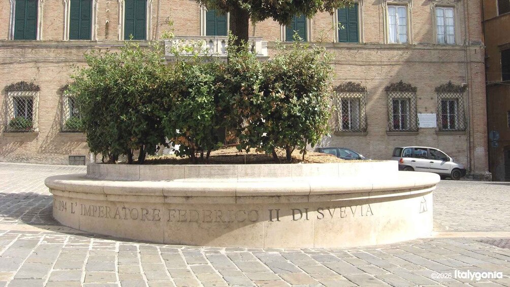 A stone fountain with inscriptions, surrounded by trees and a building with green shutters in the background. 4