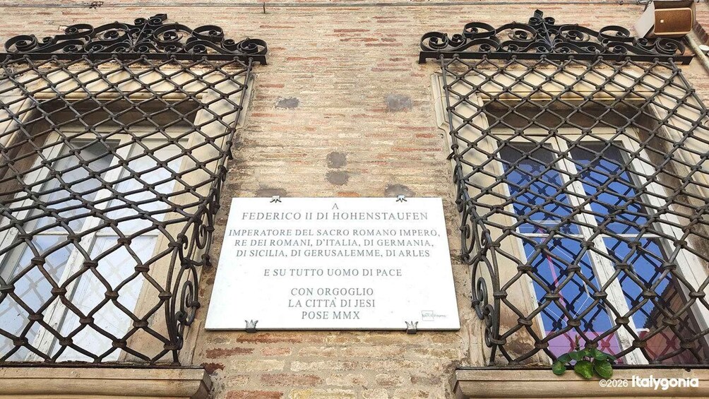 A plaque dedicated to Federico II di Hohenstaufen, with a brick wall and metal window grills in the background. 5