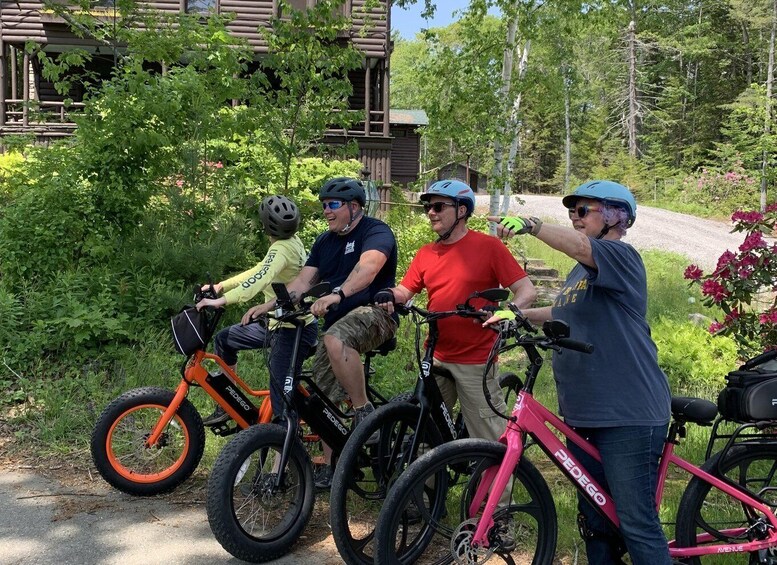 Four people on bicycles, a child and three adults, in a wooded area with a dirt path and a building in the background. 1