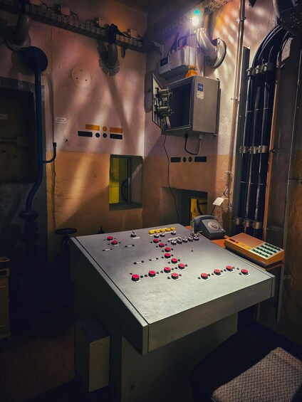 A control panel with numerous buttons and switches, a large metal table, and a complex array of pipes and ducts on the walls. 1