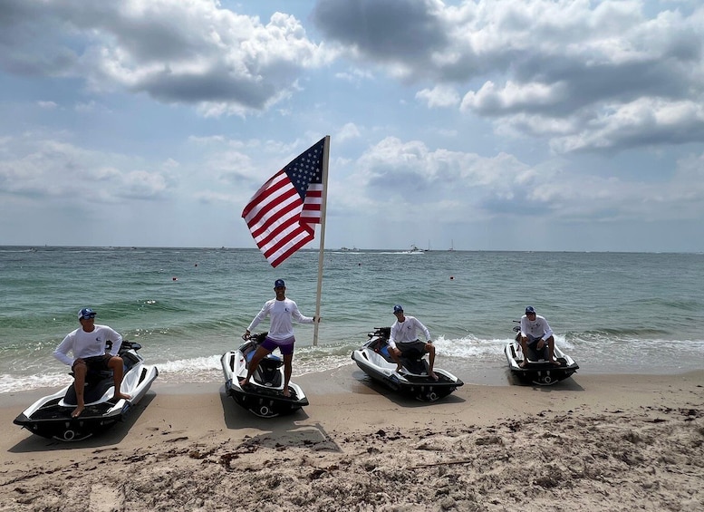 Three jet skis on a sandy beach with an American flag, a clear sky, and calm ocean waters. 3