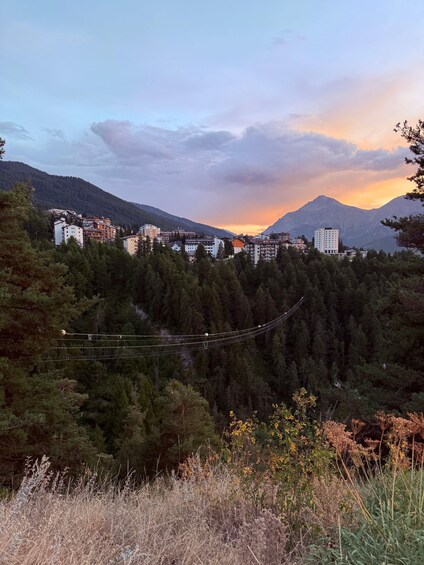 A mountainous landscape with buildings, a suspension bridge, and a sunset. 2