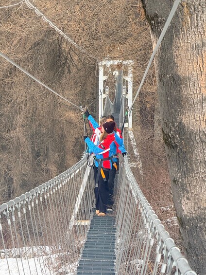 A person crossing a suspension bridge with metal railings and cables, surrounded by dry, leafless trees. 1