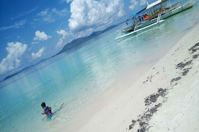 A beach with clear turquoise water, a person wading in the shallow water, and a boat with a canopy on the shore. 1