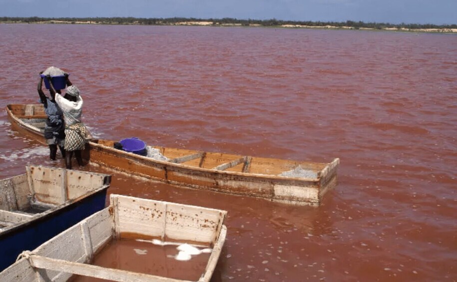 A wooden boat on a river with reddish water, a person standing in the boat, and another person in a smaller boat nearby. 5