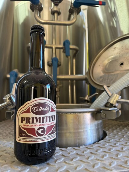A bottle of Calender Primitiva with a label, placed on a metallic surface with a valve and pipes in the background. 4