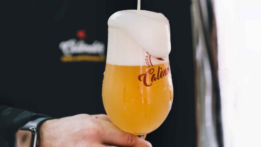A hand holding a glass of beer with a foamy head, with the word 'Caliente' visible on the glass. 1