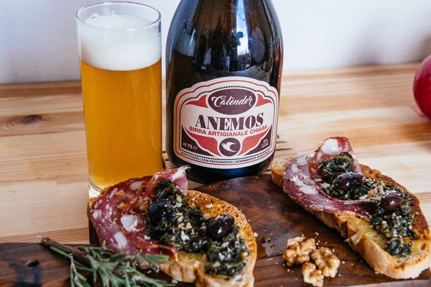 A bottle of Calender Anemos beer, a glass of beer, and two pieces of bruschetta with toppings. 3