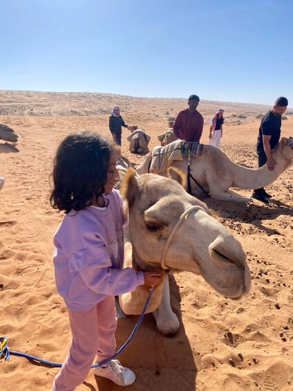 A child petting a camel in a desert with other people in the background. 3