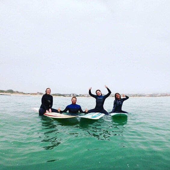 Four people sitting on surfboards in the water with a beach and buildings in the background. 4