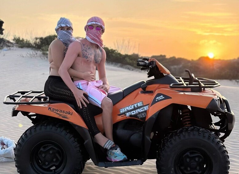 Two people on a Kawasaki ATV with 'BRUTE FORCE' written on it, sitting on a sandy beach with the sun setting in the background. 1