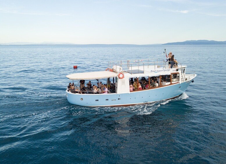 A light blue boat with 'Boat tours Opatija' written on the side, sailing on calm blue waters with a clear sky and distant mountains in the background. 2
