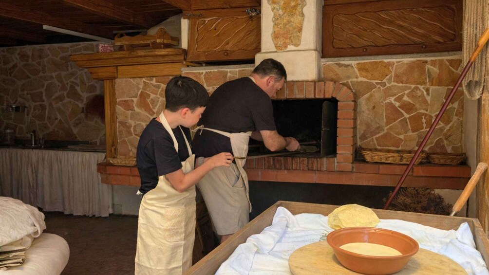 Two individuals in a rustic kitchen with a stone wall and wooden beams, preparing food in a wood-fired oven. 5