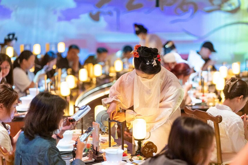 A traditional tea ceremony with participants seated at tables, a tea server in the center, and a decorative backdrop. 1