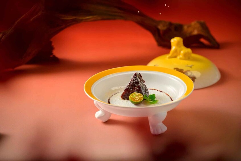 A dessert on a small white stand with a yellow rim, garnished with a mint leaf and a slice of lime, set against a red background with a decorative element. 5