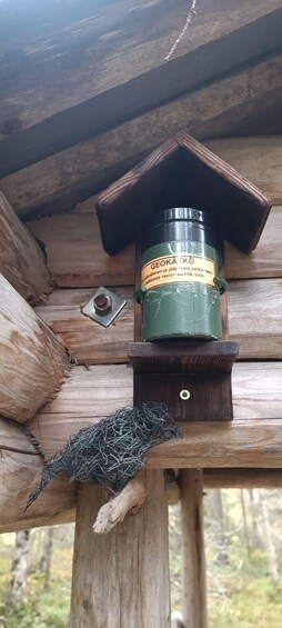 A wooden birdhouse with a green container labeled 'GEOKATKO' on top, a metal latch, and a nest with twigs at the base. 3