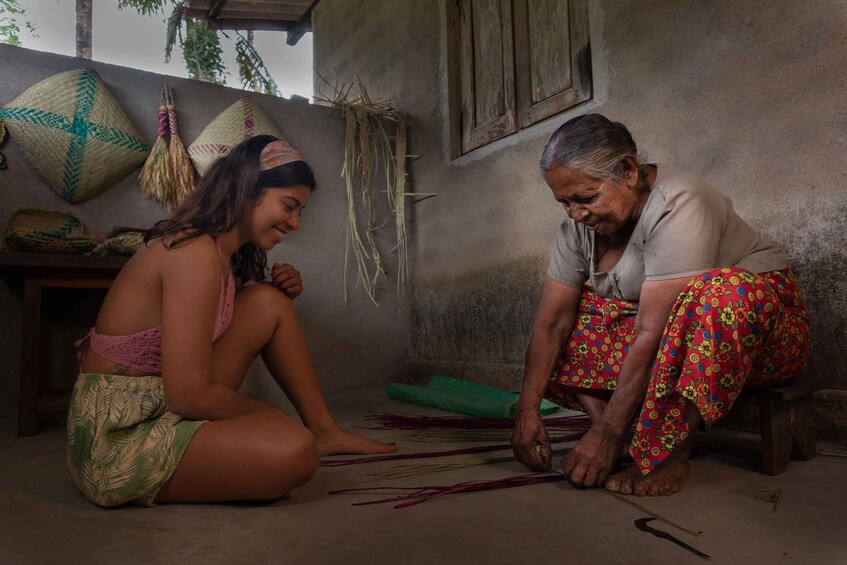 Two people sitting on the floor, one wearing a patterned skirt and the other in a floral skirt, with woven baskets and a wall in the background. 3